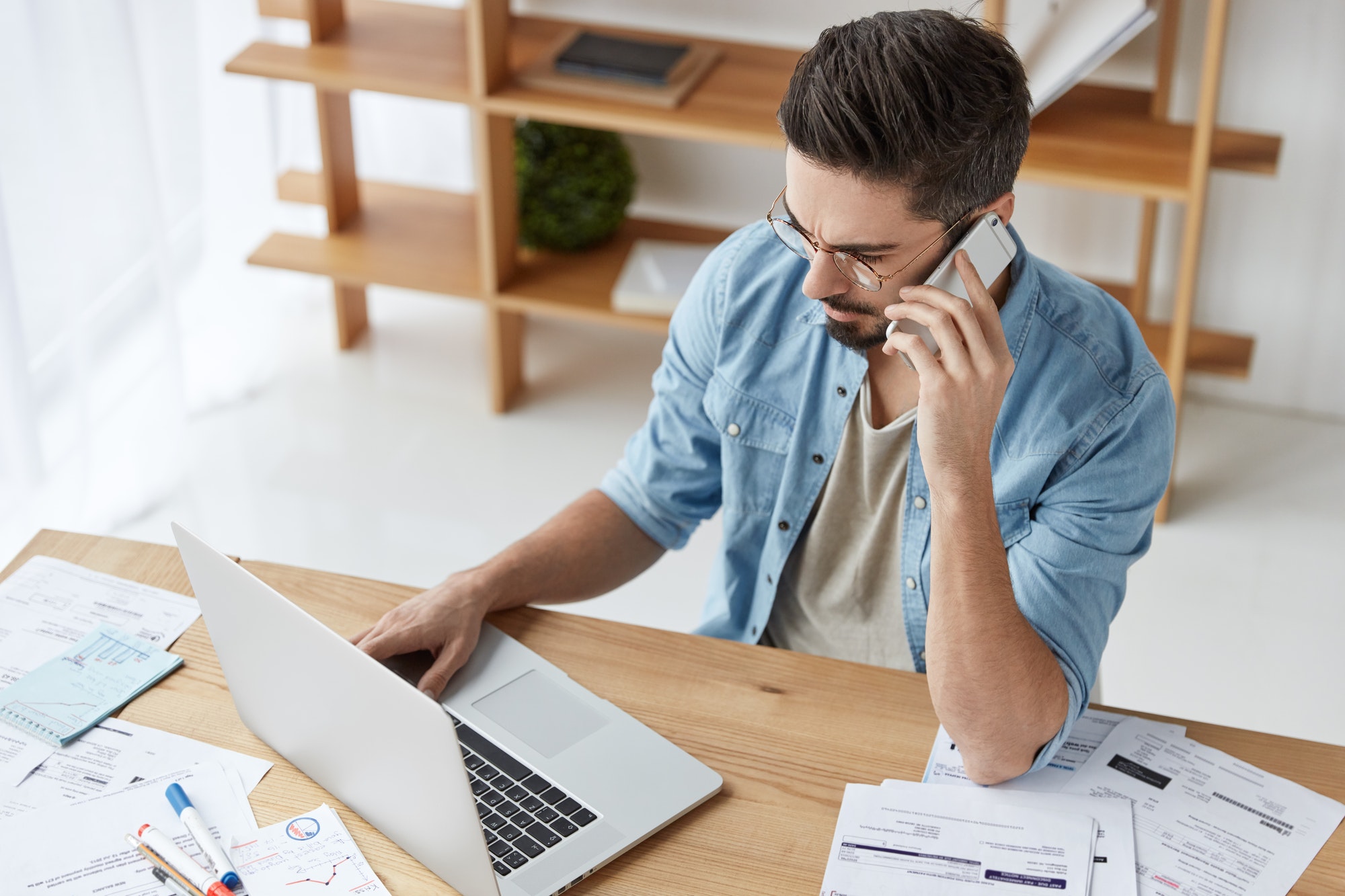 A man talking to someone on his phone while using a laptop and some paper on his table